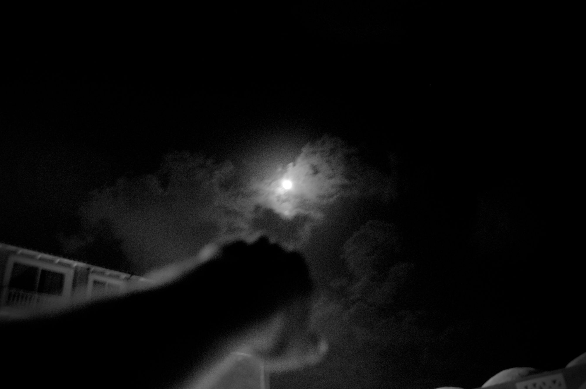 A black and white photo of someone pointing a fist at the moon behind clouds.